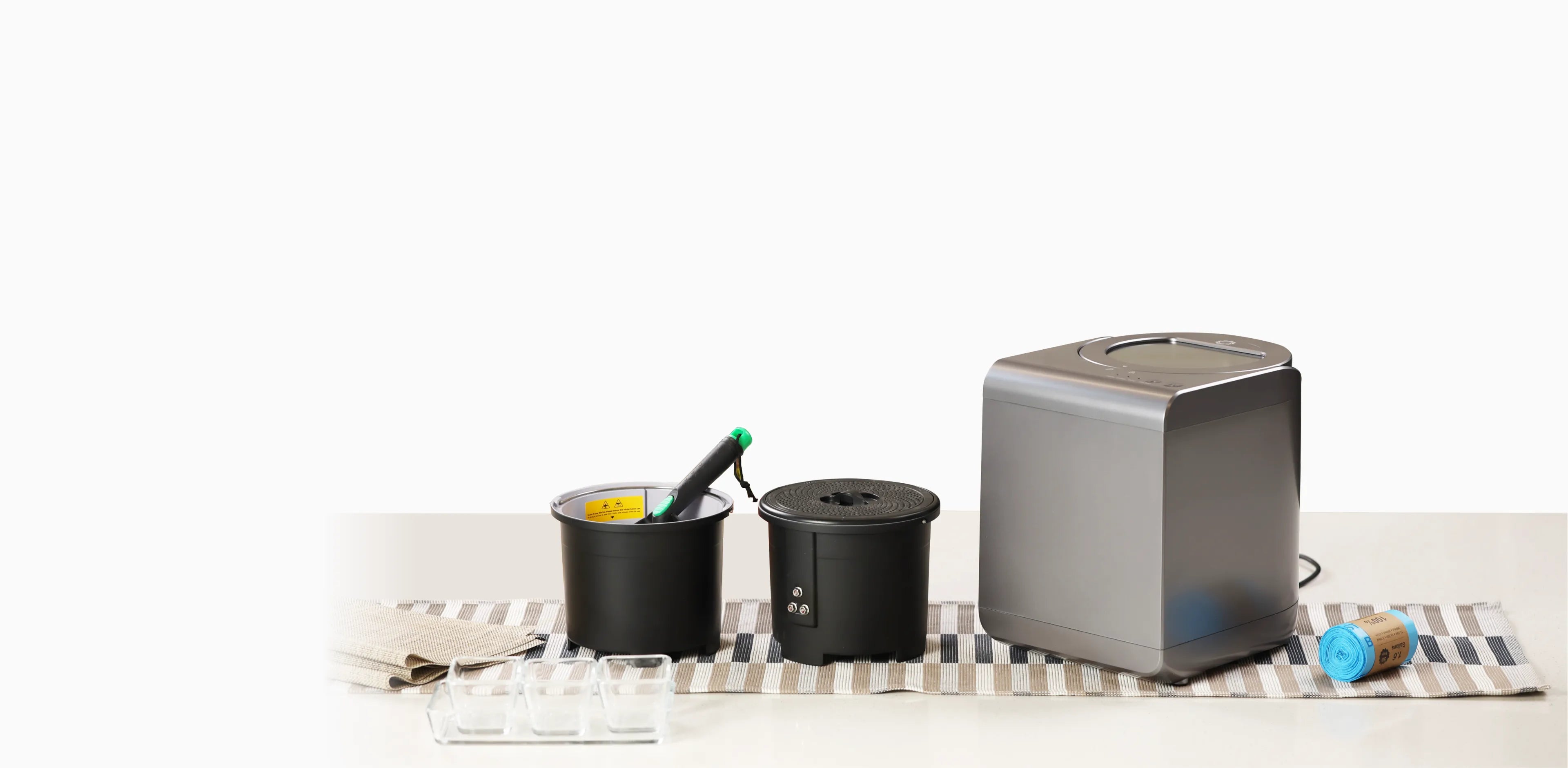 カウンタートップのセットアップには、フードプロセッサー、緑のハンドルが付いた黒い容器2つ、青いプラスチックラップのロールが含まれています。これらのアイテムは、ストライプのキッチンタオルの上に置かれています。