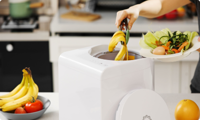 キッチンで、白いAirtherealコンポスト機械にバナナの皮や野菜くずを追加している人