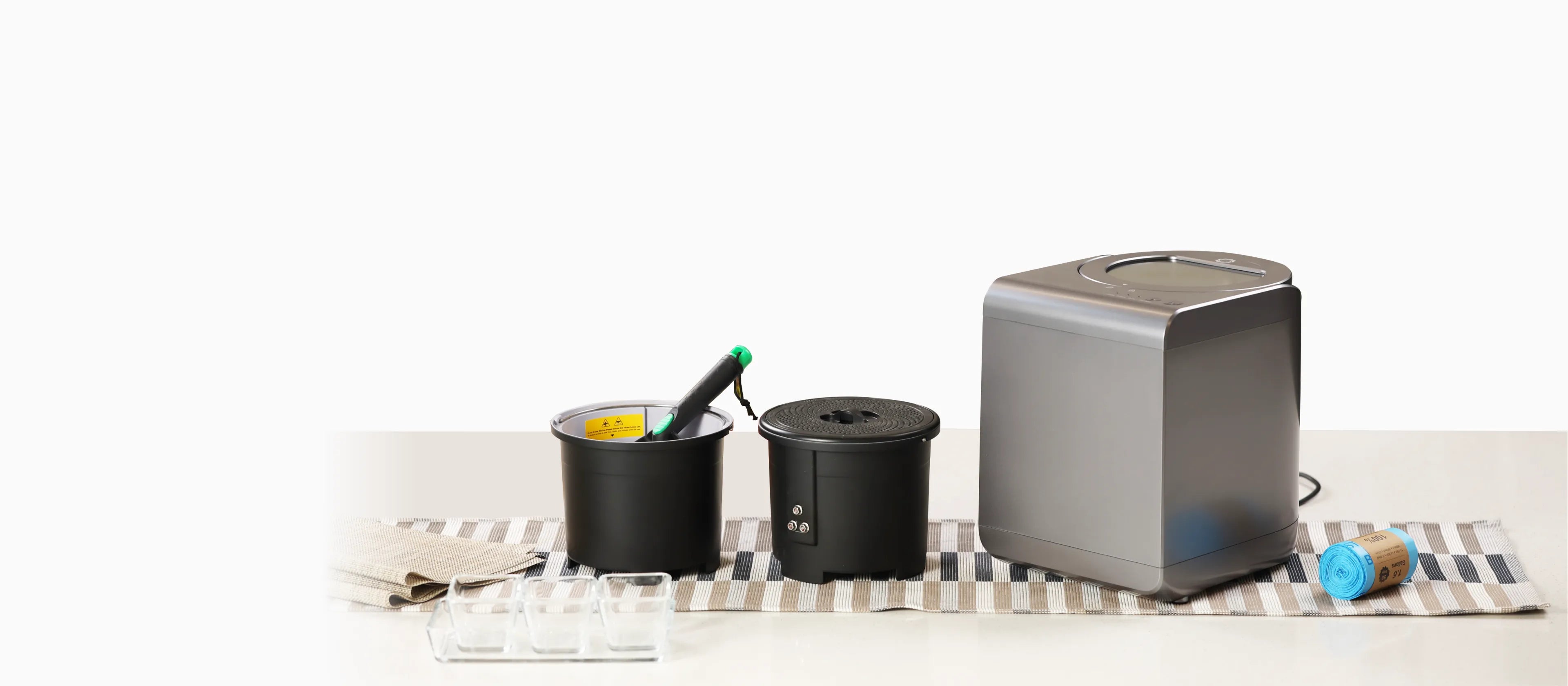 カウンタートップのセットアップには、フードプロセッサー、緑のハンドルが付いた黒い容器2つ、青いプラスチックラップのロールが含まれています。これらのアイテムは、ストライプのキッチンタオルの上に置かれています。