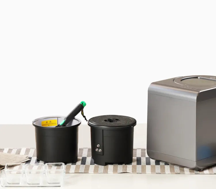 カウンタートップのセットアップには、フードプロセッサー、緑のハンドルが付いた黒い容器2つ、青いプラスチックラップのロールが含まれています。これらのアイテムは、ストライプのキッチンタオルの上に置かれています。